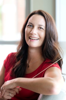 Brunette woman wearing a red dress and holding a baton. She is seated and smiling at the camera.