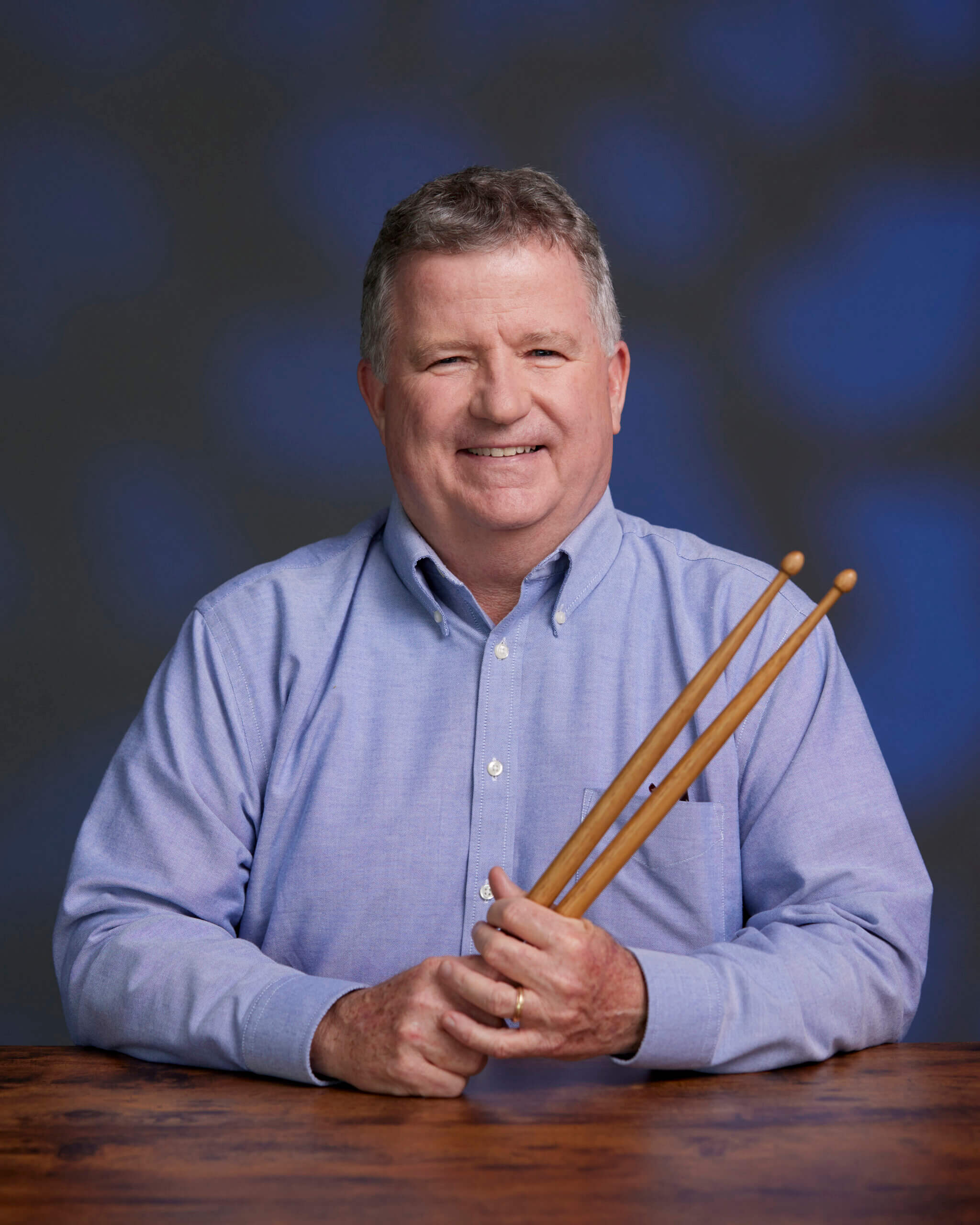 Man in long-sleeved light blue shirt holding pair of drumsticks.