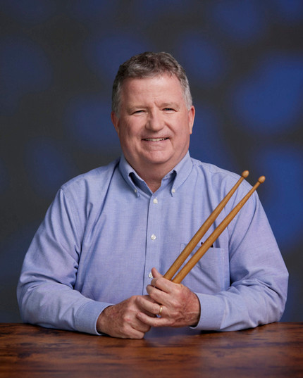 Man in long-sleeved light blue shirt holding pair of drumsticks.