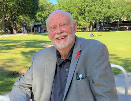 Older man with short gray hair and wearing a gray suit, sitting near the UNF green on a bright, sunny evening and smiling.