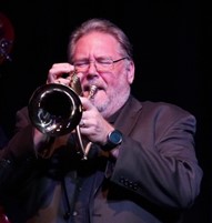 Man in black suit blowing a trumpet. 