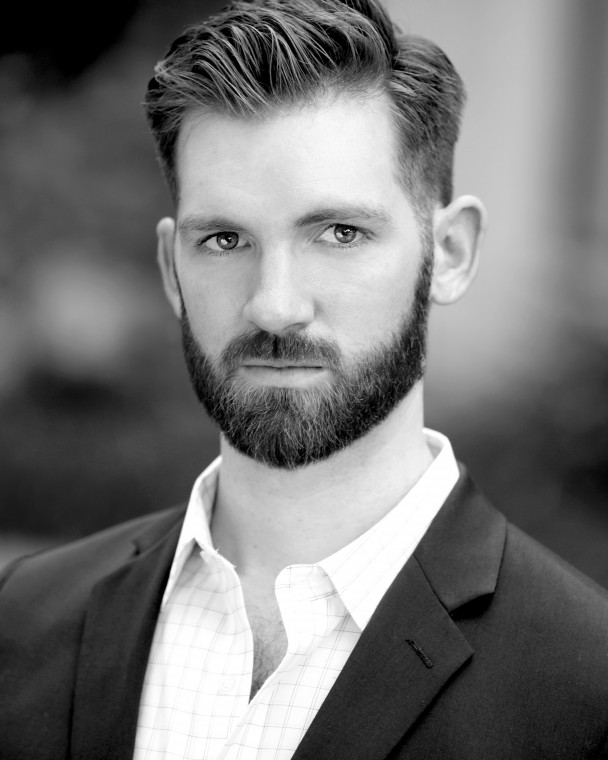Black and white headshot of Caucasian male with full beard. He is wearing a dark blazer with a white collard shirt. 
