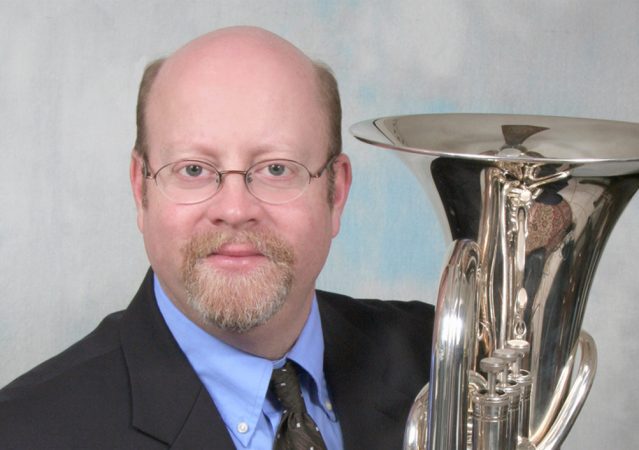 Man with light brown hair and goatee. He wears glasses and is holding a tuba. 