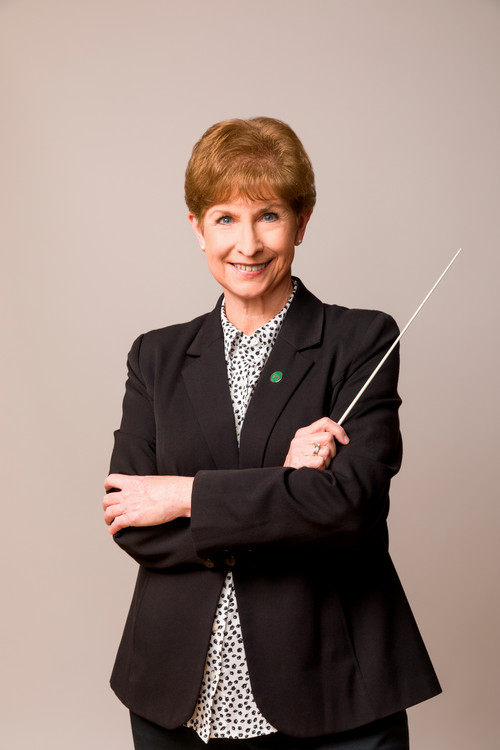 A smiling female conductor in a black blazer, holding a conductor's baton, exuding confidence and approachability.