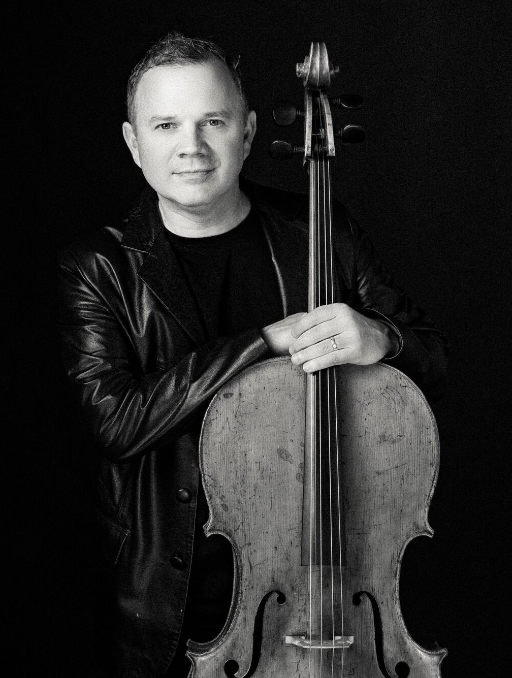 Black and white photo of white male dressed in all black and leaning against neck of cello. 