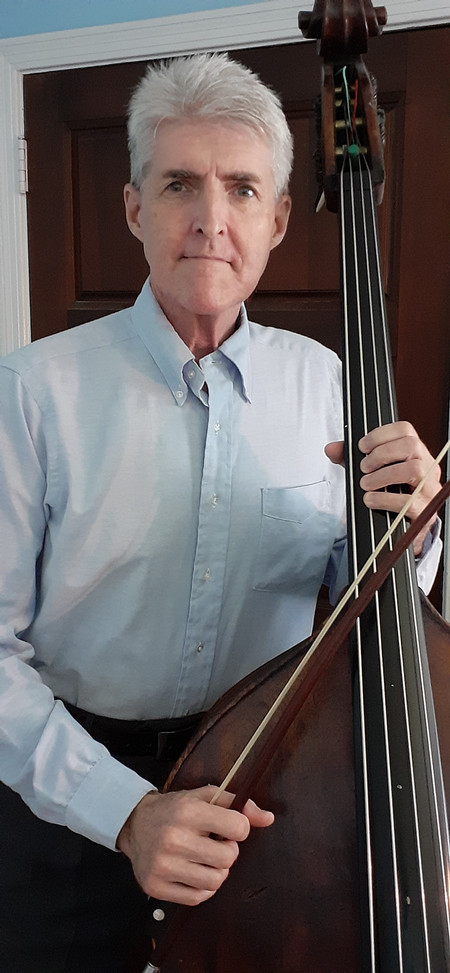 Man with white hair and long sleeved white shirt, standing and holding double bass. 