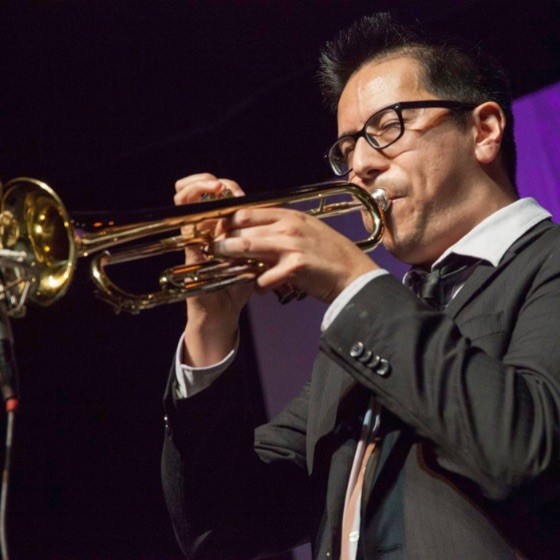 Man wearing black framed glasses and blowing a trumpet. 