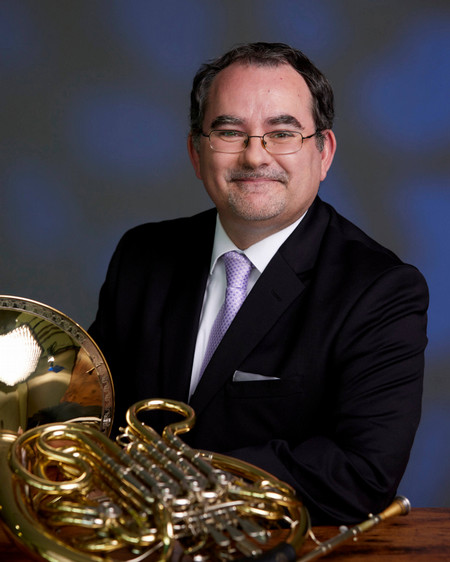 Man with short black hair wearing dark suit. His French horn is resting on the table in front of him.