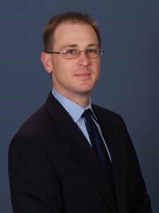 Man with short brown hair wearing glasses and dark blue blazer with light blue collard shirt. 