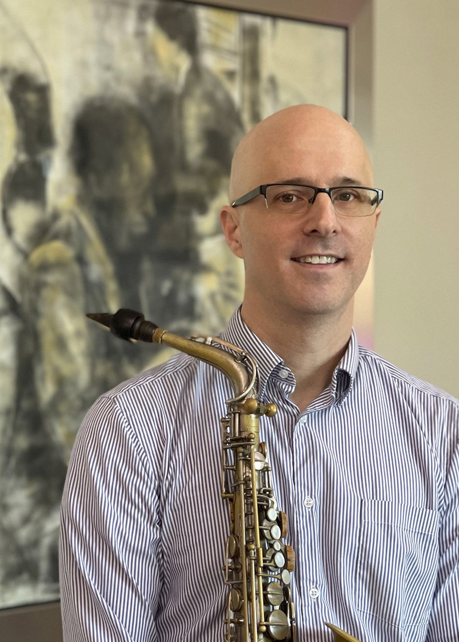 Man wearing glasses and holding saxophone. He wears a striped navy collard shirt. 