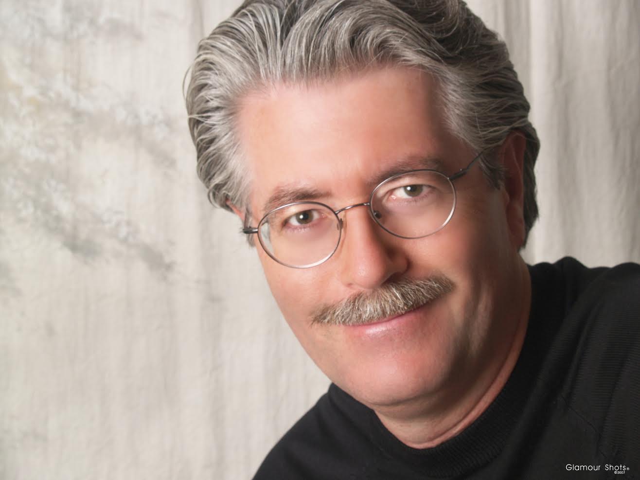 Man with silver hair and moustache wearing thin-framed silver glasses.