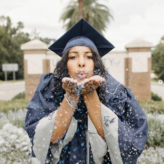 UNF female student wearing a cap and gown and blowing confetti at the camera