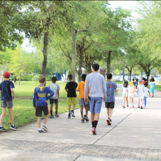 A photograph of campers walking to the UNF library from COEHS. It is a bright day and the trees are a vibrant green.