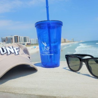 UNF merch such as a hat, tumbler, and glasses on a pier at the beach