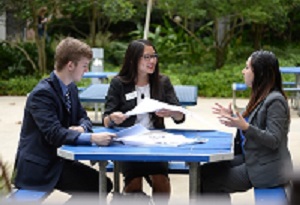 3 students sitting at a blue picnic table talking with notebooks