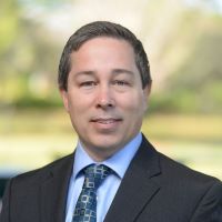 Headshot of Doug Titus with a suit and blue tie with a green blurred background