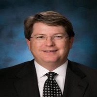 Headshot of Nelson Bradshaw wearing a suit and checkered tie with blue and black background