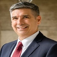 Headshot of Sam Inman wearing suit and red tie with a blurred gold background