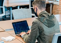 A male student wearing a mask and coding on a laptop with back toward camera