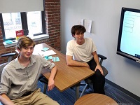 Two male students in a study room sitting at a table with window, brick wall and tv