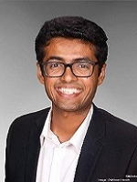 Headshot of Riju Thomas wearing suit and glasses with gray background