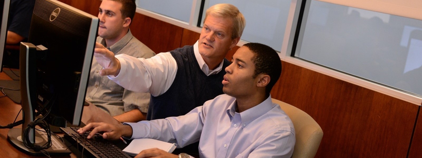 A professor standing by two students on the computer, while talking and pointing at the computer screen