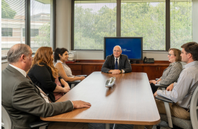 Six people wearing business attire sitting at a conference table with windows and a TV in the background