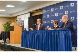 The dean speaking at the podium with 3 people sitting at a desk speaking through microphone