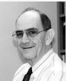 A black and white headshot of a man with a collared shirt and tie wearing glasses