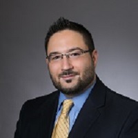 Headshot of Justin Riddell wearing a suit with  yellow tie, glasses and a gray background.