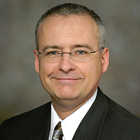Headshot of Rich Goeldner wearing a suit and tie with a gray background.