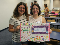 Two people wearing glasses posing with a multicolored sign with desks and people in the background