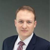 Headshot of jonathan Mann wearing suit with red striped tie with gray background