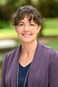 Headshot of Kate Learch wearing a purple blazer with a blue shirt with a green blurred background