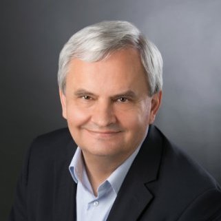 Headshot of Greg wearing a black blazer and blue collared shirt with gray background