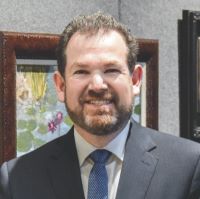 Headshot of Adam Chaskin wearing suit with blue tie with a framed picture behind him