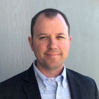 Headshot of Casey Wagner wearing black blazer with light blue shirt and gray background