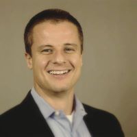 Headshot of Benjamin H wearing black blazer and blue collared shirt with brown background