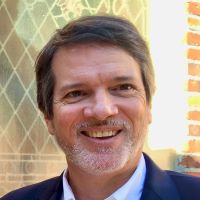 Headshot of Drew T wearing blue blazer and white collared shirt with brick background