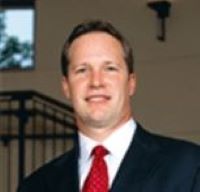 Headshot of Gregory Gundlach wearing suit and red tie with window and stair railings in background