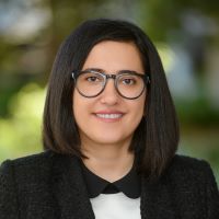 Headshot of Hanieh S wearing black blazer and white and black shirt with glasses and green blurred background