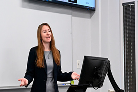 Female professor in black blazer with grey undershirt lecturing by a whiteboard and computer monitor 