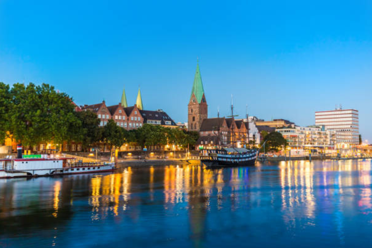 Trees, boats and old style European buildings sit underneath a blue sky aligned along a reflective riverfront