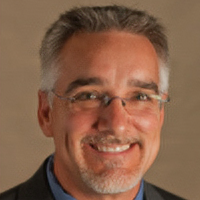 Headshot of Scott Lane with glasses, a black blazer, blue shirt and tie with a brown background