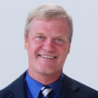 Headshot of David Swenson wearing black blazer, blue shirt and striped tie with gray background