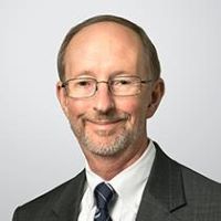 Headshot of Paul Hitchcock wearing a suit, black tie and glasses with gray background