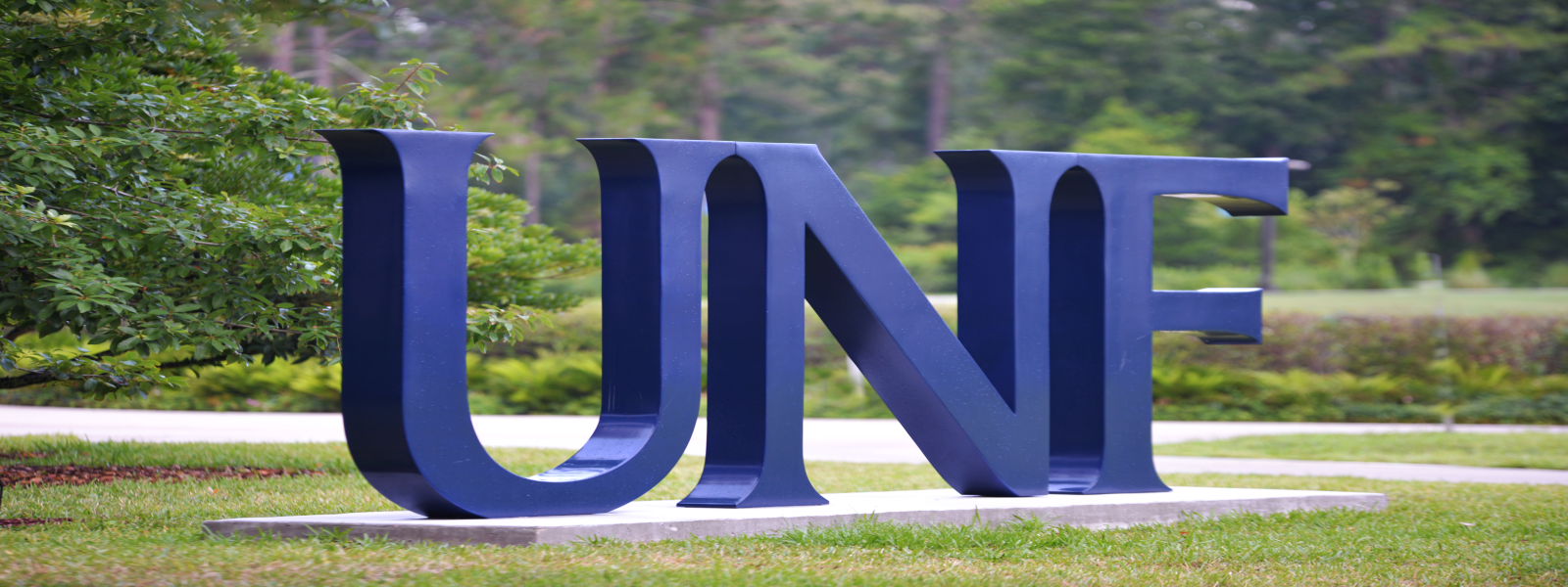 A photo of a sign with a blue letter U, N and F with trees, bushes and a sidewalk in the background.