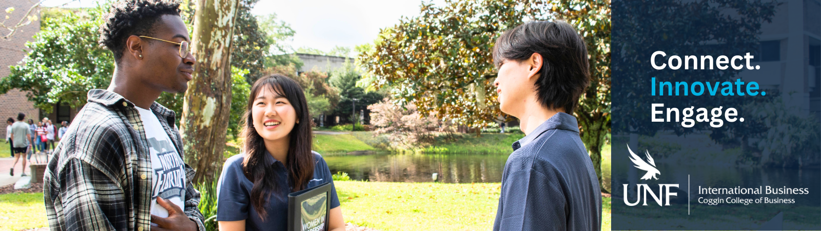 three international Coggin students talking outside in front of the lake and buildings on campus