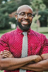 Headshot of Sergio Kelsor wearing a red shirt with white dots, a gray tie with blurred tree background