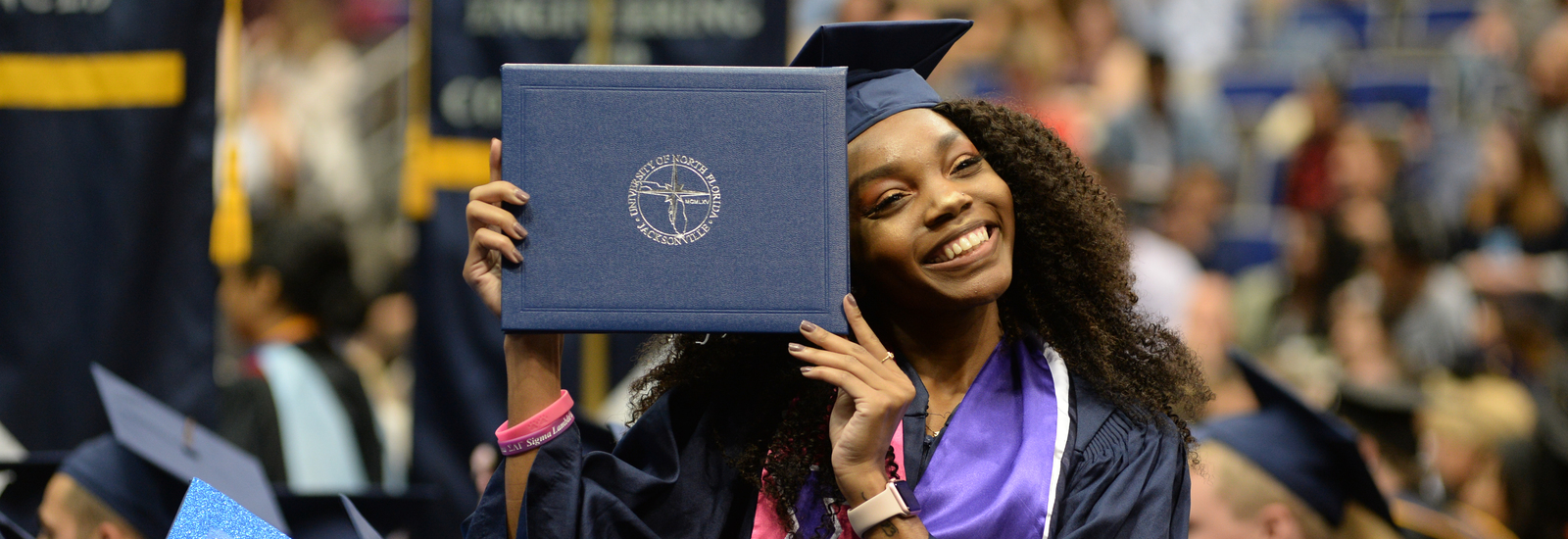 Female student graduating holding up a UNF diploma cover by her face and smiling